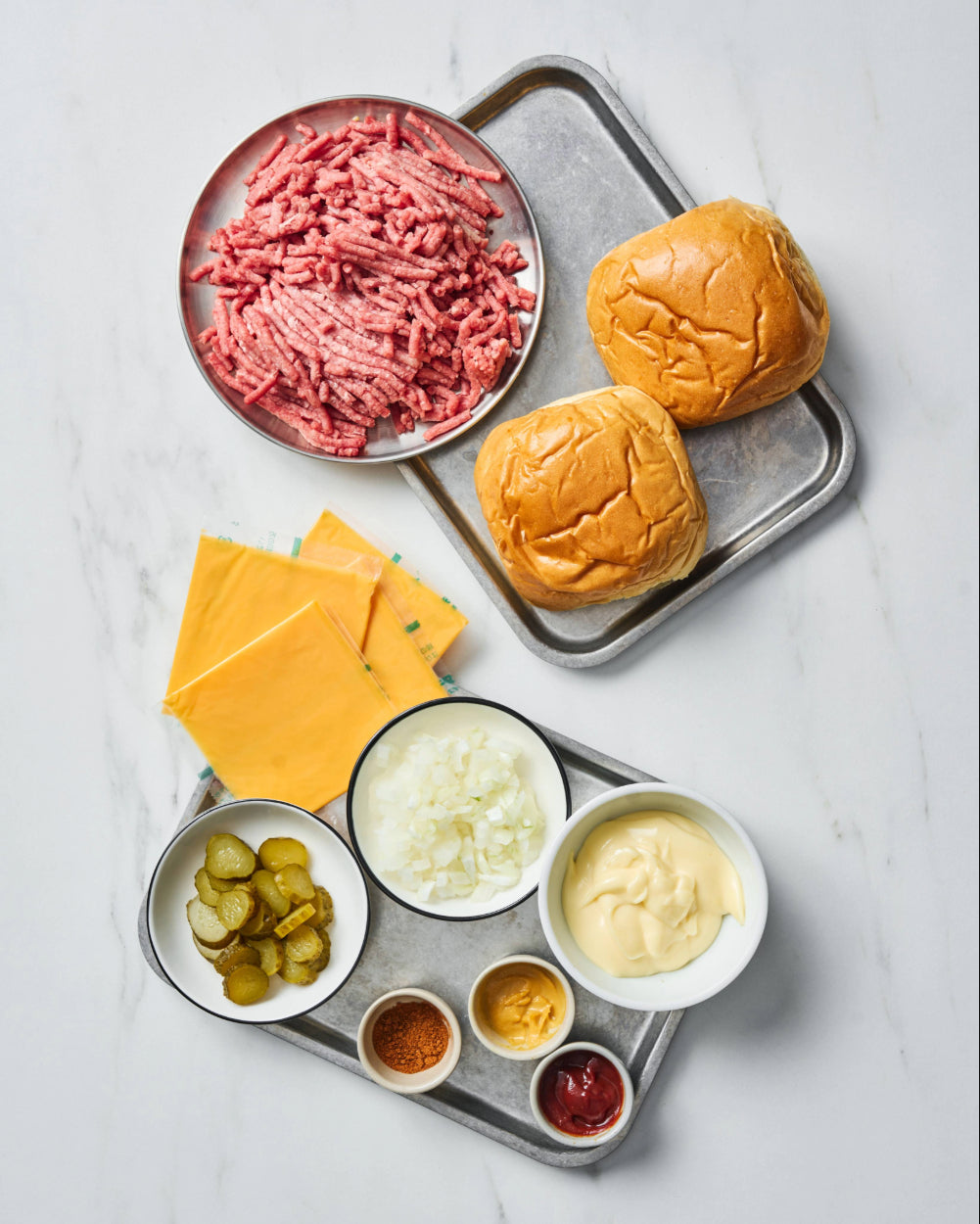 Smash burger ingredients laid out on a marble surface including ground beef, brioche buns, American cheese slices, pickles, diced onion, mayonnaise, mustard, ketchup, and paprika