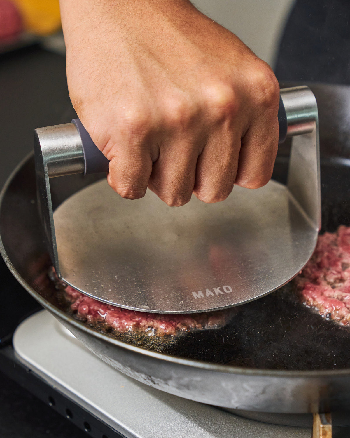 A MAKO press smashing a beef patty flat in a hot MAKO carbon steel steak pan on a stovetop