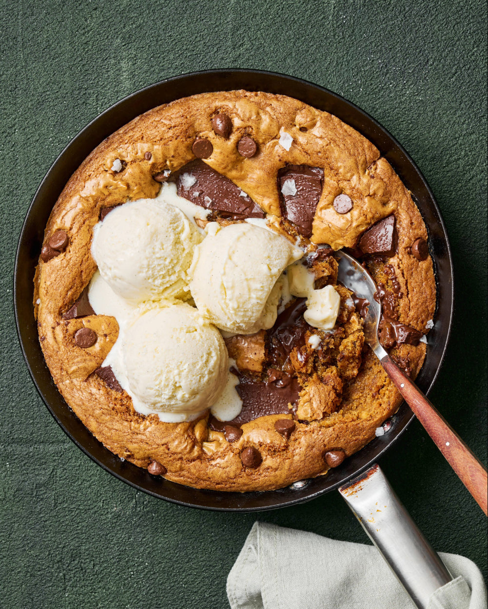 A baked chocolate chip skillet cookie topped with three scoops of vanilla ice cream, with a spoon scooping into the gooey center to reveal melted chocolate.