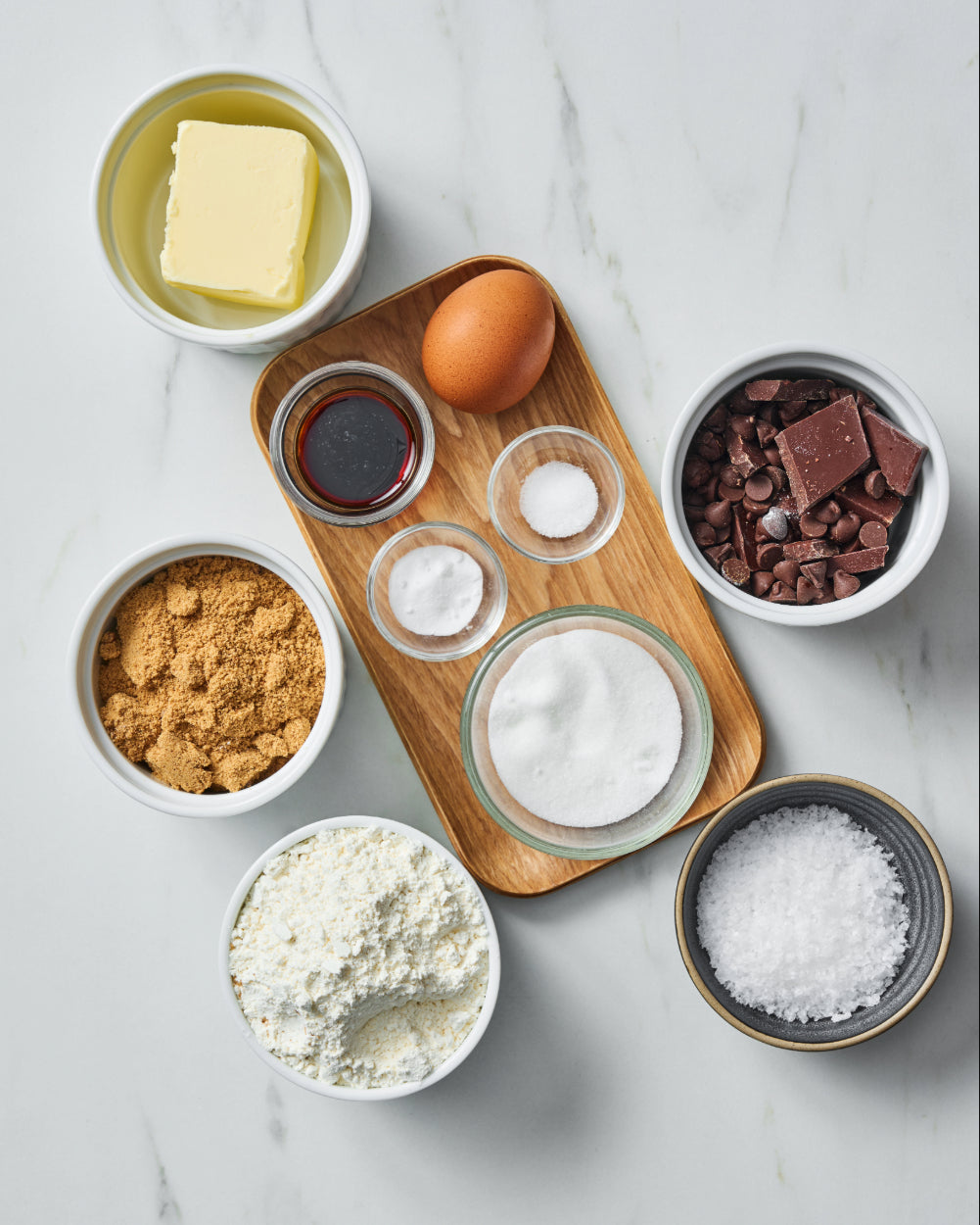 Overhead view of skillet cookie ingredients - butter, a brown egg, vanilla extract, baking soda, white sugar, brown sugar, flour, chocolate chunks and chips, and flaky sea salt.
