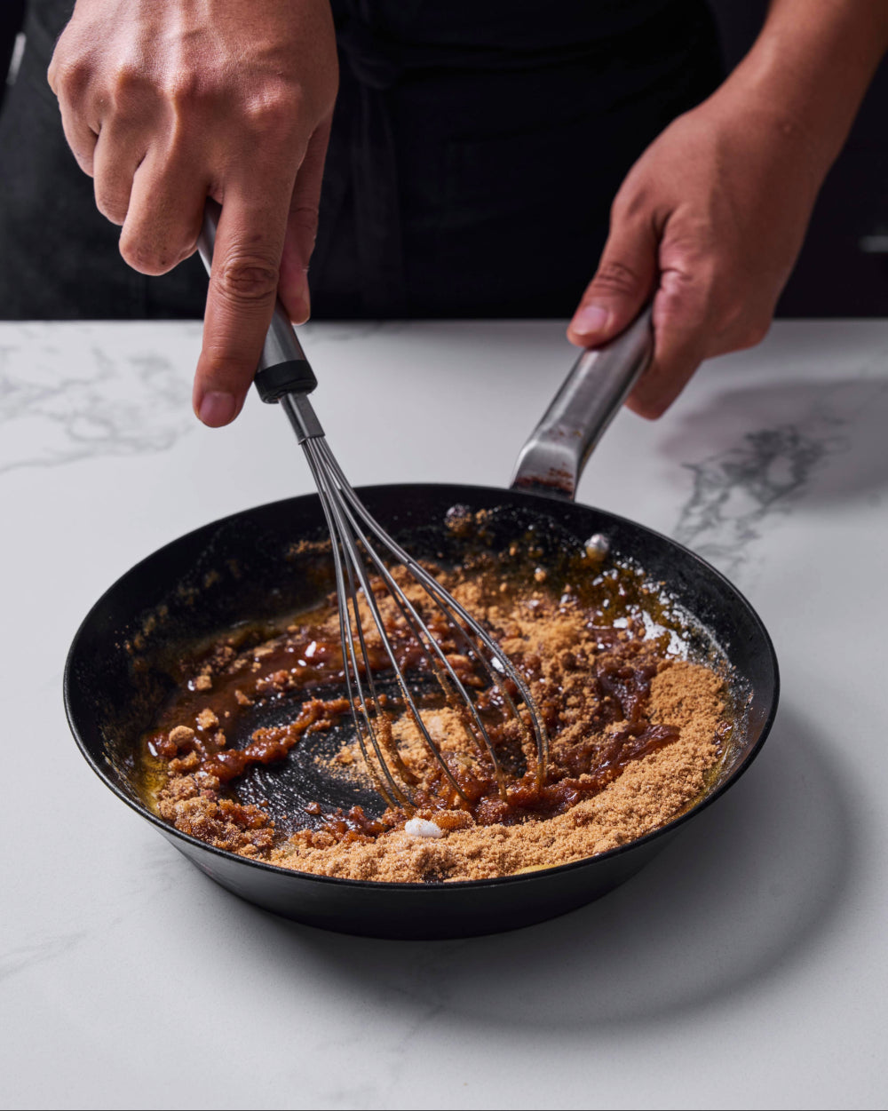 Whisking brown sugar into melted butter in a black skillet on a marble countertop, blending the ingredients into a glossy caramel-colored mixture.