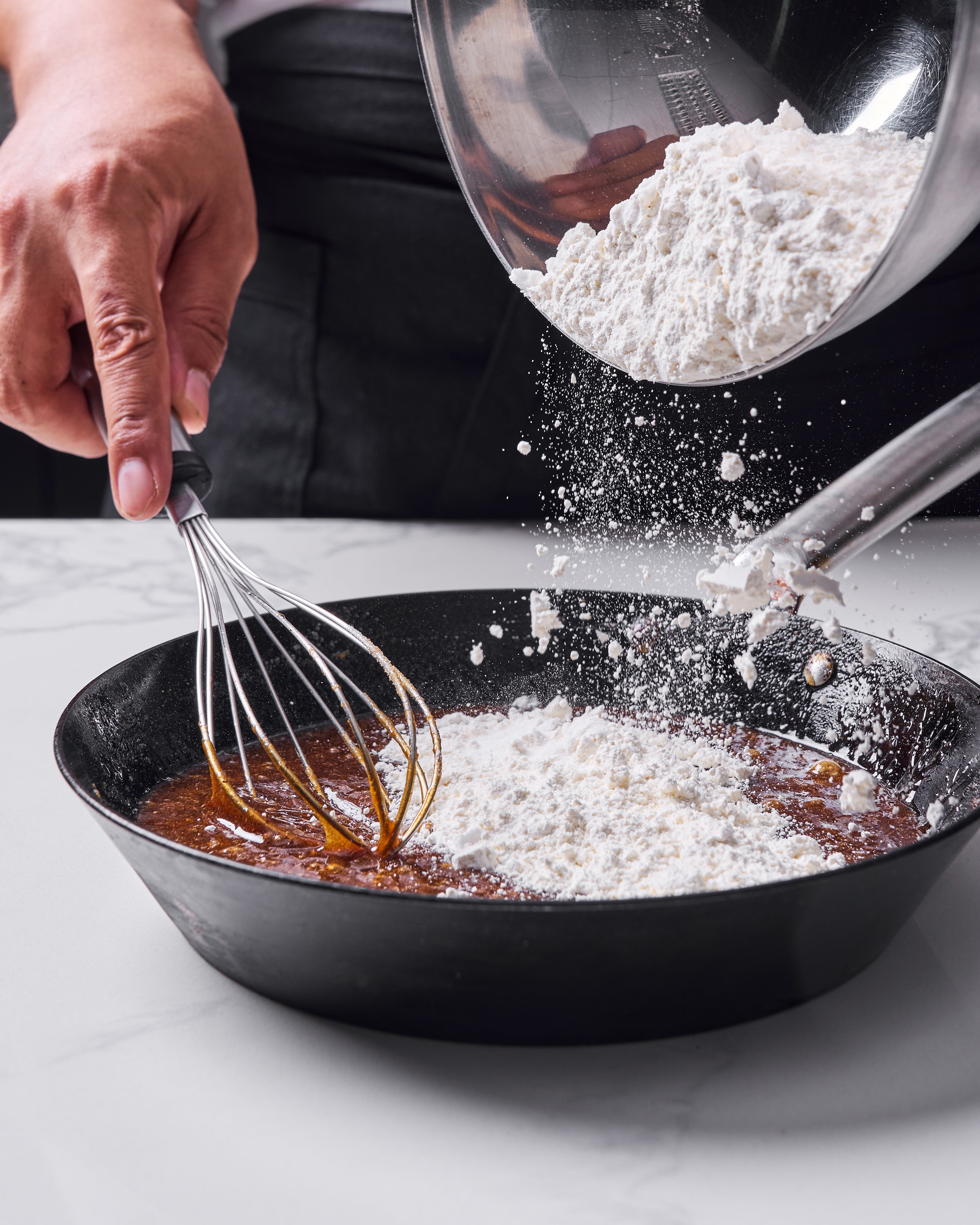 Flour being poured from a metal bowl into a black skillet containing a glossy caramel-colored batter, with a hand holding a whisk ready to mix the ingredients together.