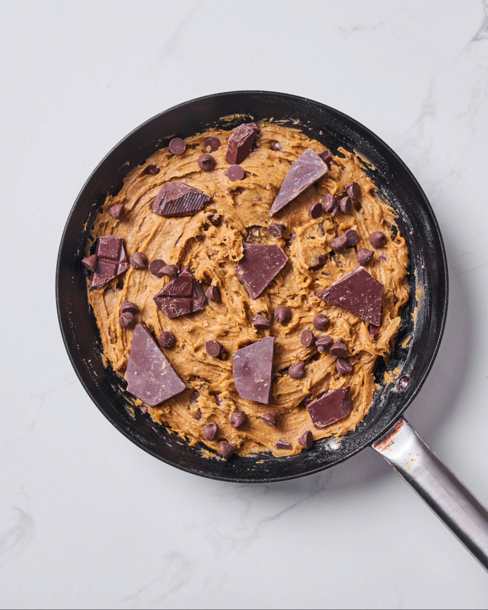 Unbaked cookie dough spread evenly in a black skillet, topped with chocolate chips and large chunks of dark chocolate, ready to go into the oven.