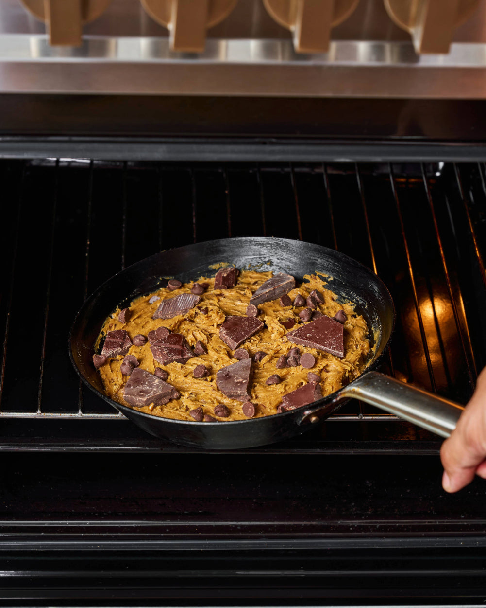A hand sliding a black skillet filled with raw chocolate chip cookie dough into an oven, ready to bake.