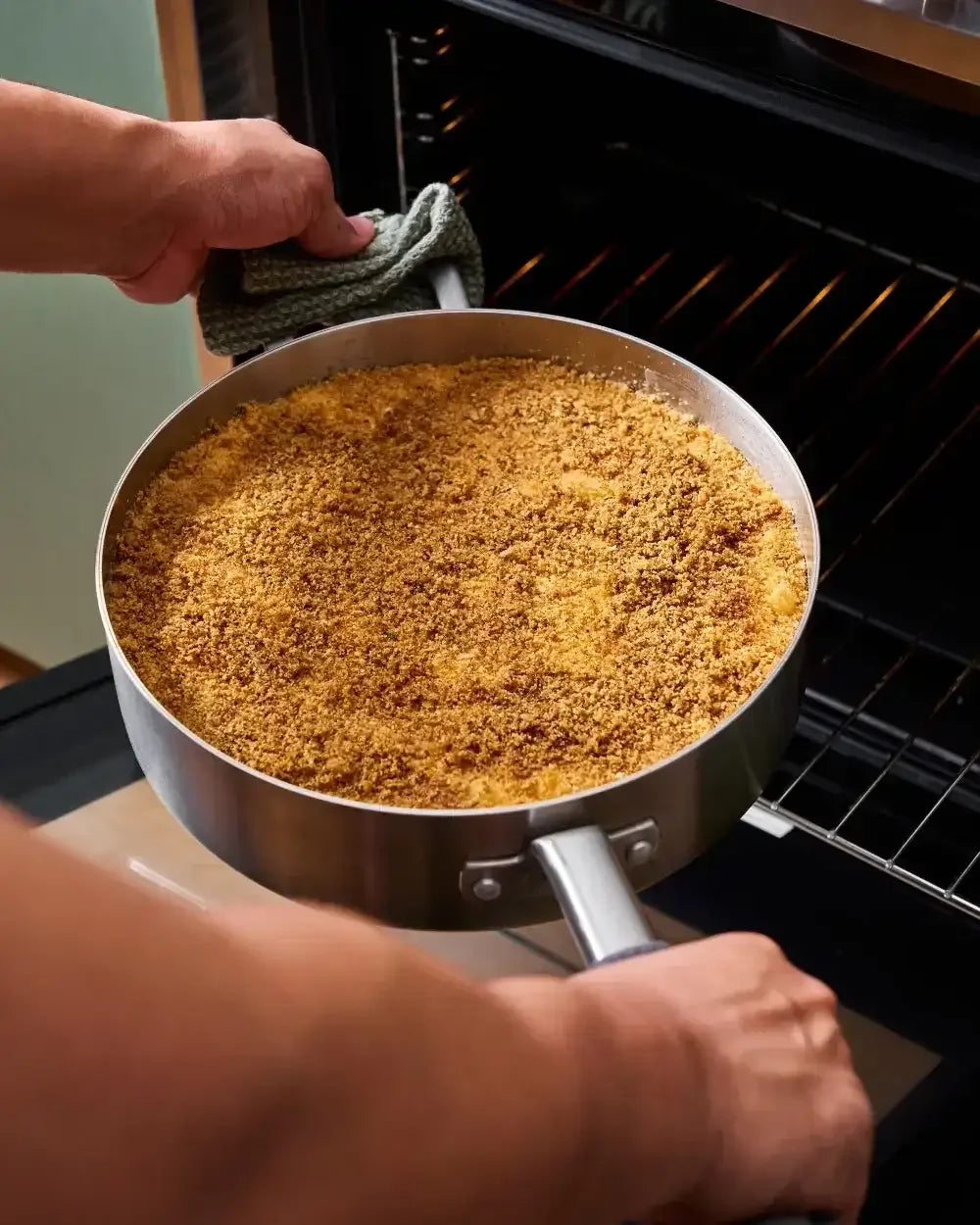 Person placing a stainless steel sauté pan with baked crumb topping into an oven