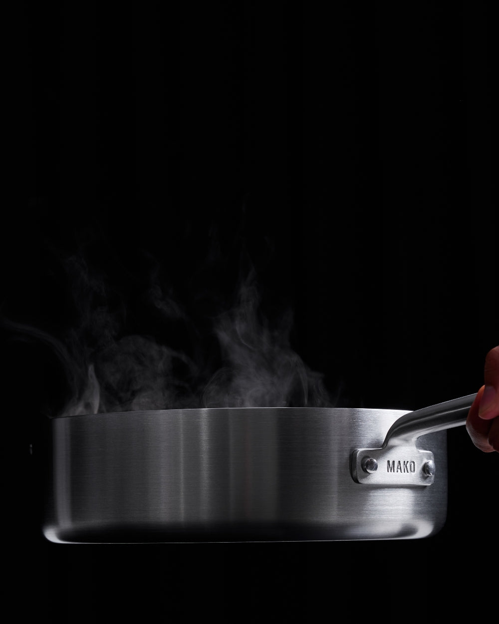 Stainless steel sauté pan with steam rising, held by hand against dark background