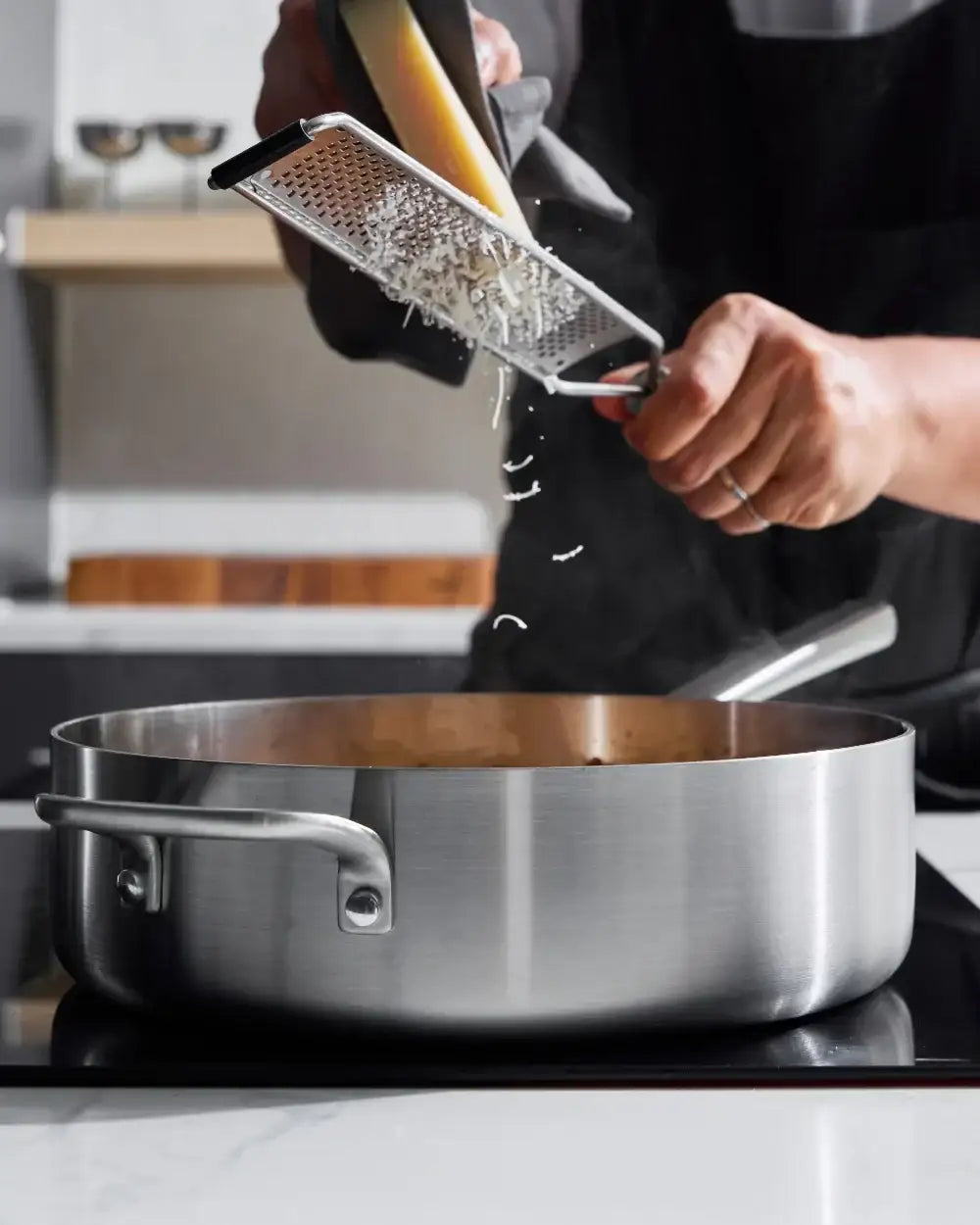 Hands grating cheese over a steaming stainless steel Mako saute pan in a modern kitchen