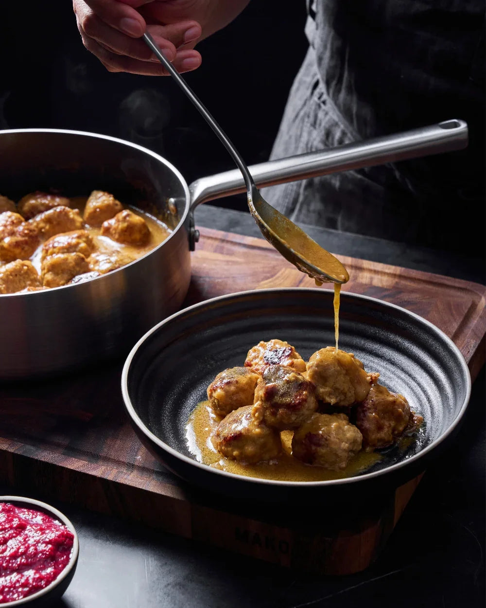 Hand pouring golden sauce over meatballs in black bowl beside stainless steel sautepan on wooden board