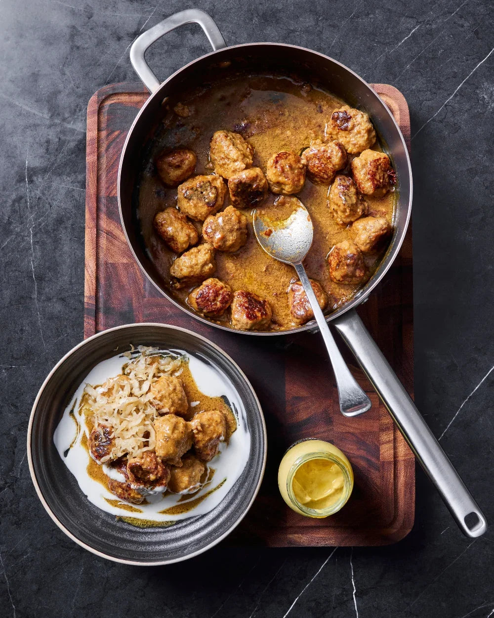 Mako saute pans cooking browned meatballs in sauce with bowl of meatballs and mustard jar on wooden board
