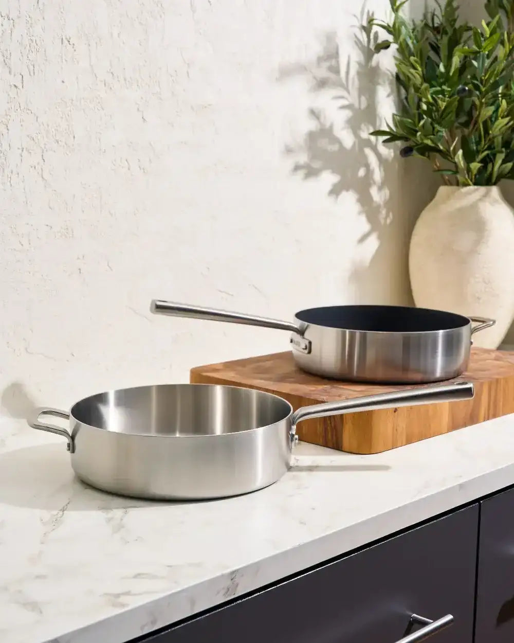 Stainless steel Mako saute pans on marble kitchen countertop with wooden board and green plant vase