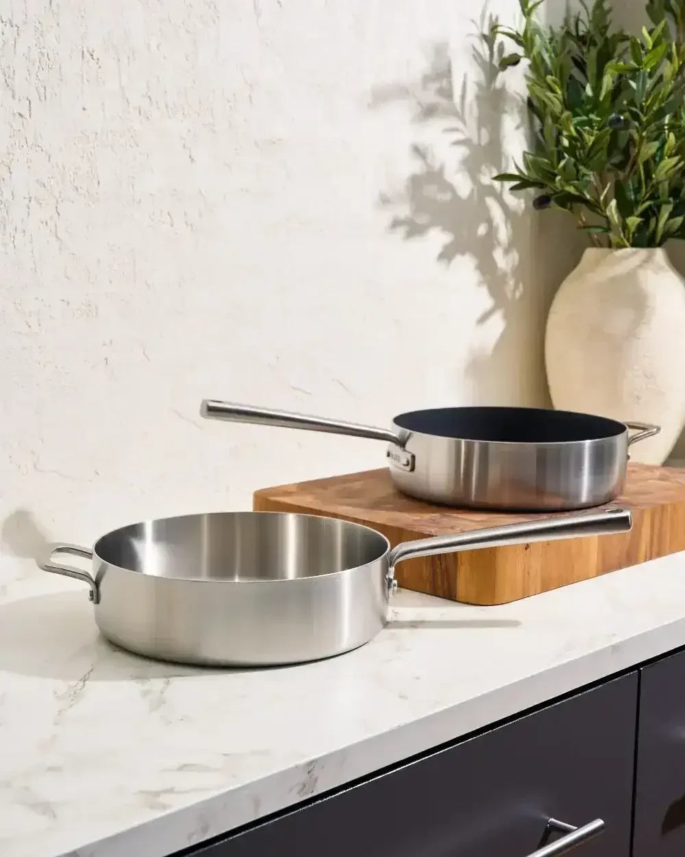 Stainless steel sauté pans with handles on a marble kitchen countertop and wood cutting board