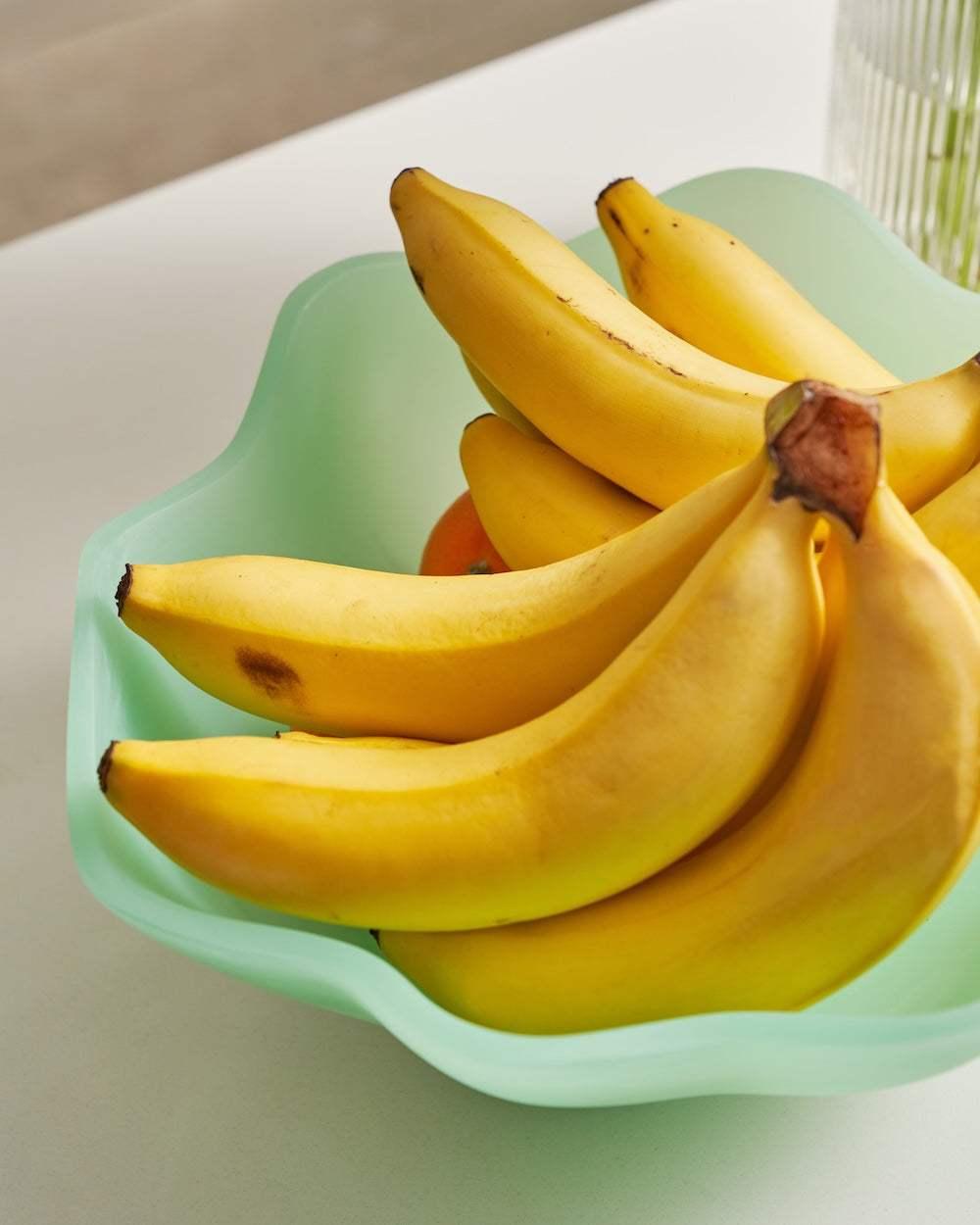 Fresh yellow bananas in a light green wavy bowl on a white table, minimalist kitchen scene