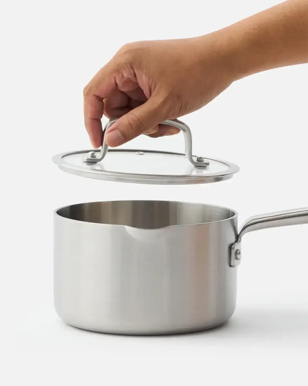 Hand lifting lid from stainless steel saucepan with handle on white background.