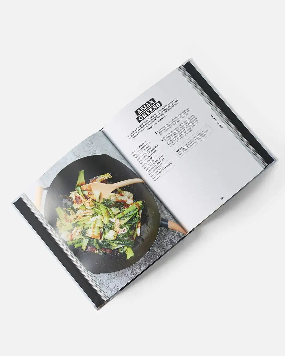Open cookbook by Marion Grasby showing Asian Greens recipe and stir-fried greens in a wok with wooden spoon