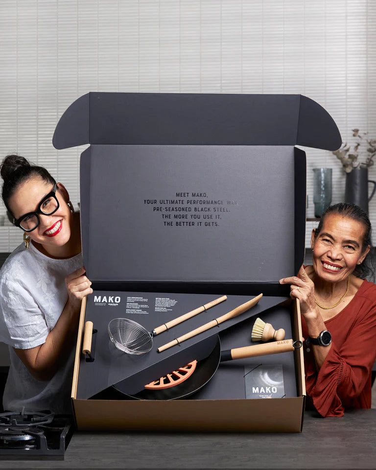 Two women presenting a MAKO cookware set in a gift box, featuring a wok and kitchen utensils.