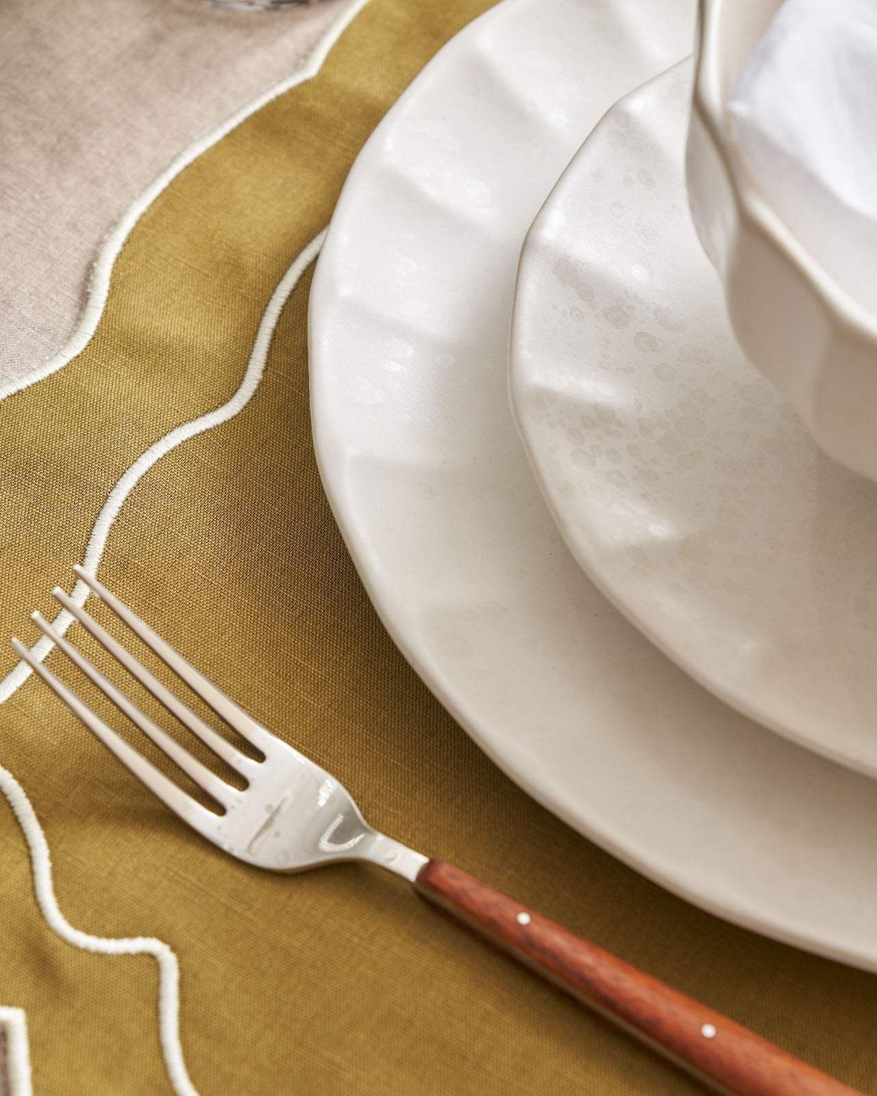 White ceramic plates with textured edges and wooden-handled fork on olive green and beige embroidered tablecloth