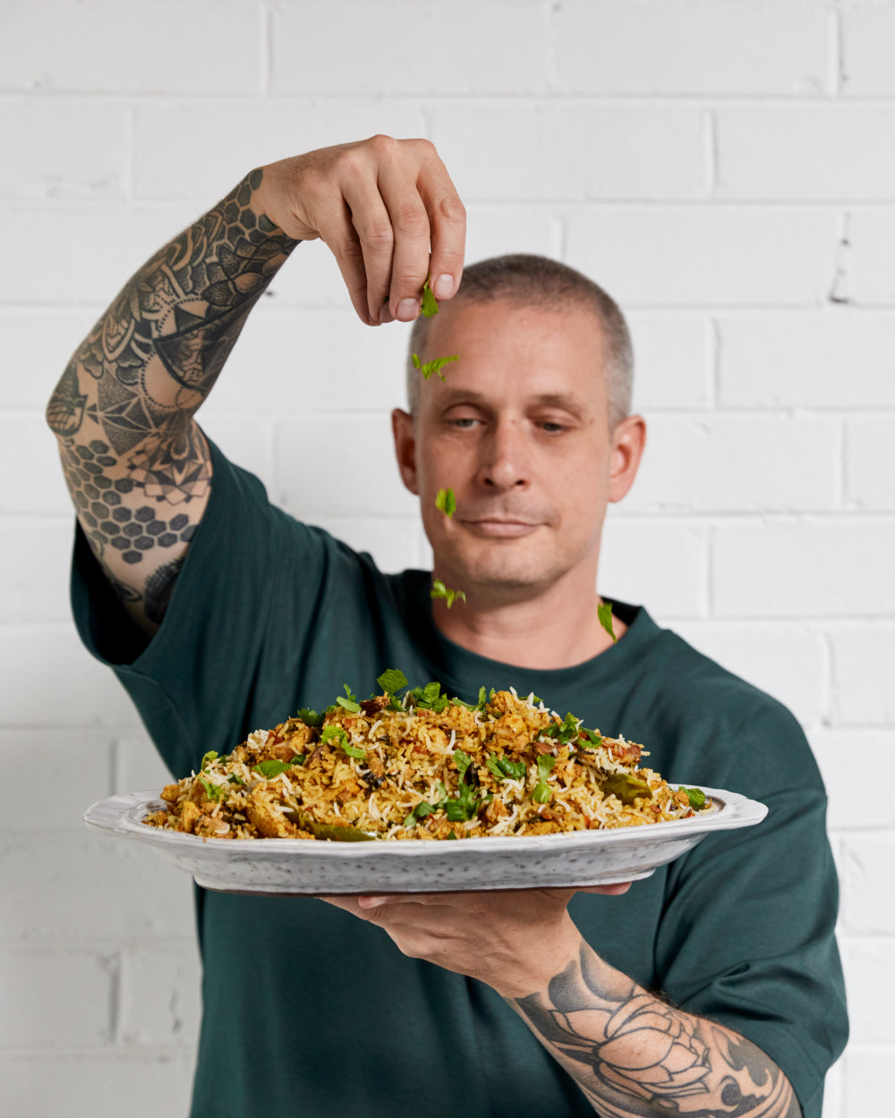 Tattooed chef garnishing rice dish with herbs in front of white brick wall