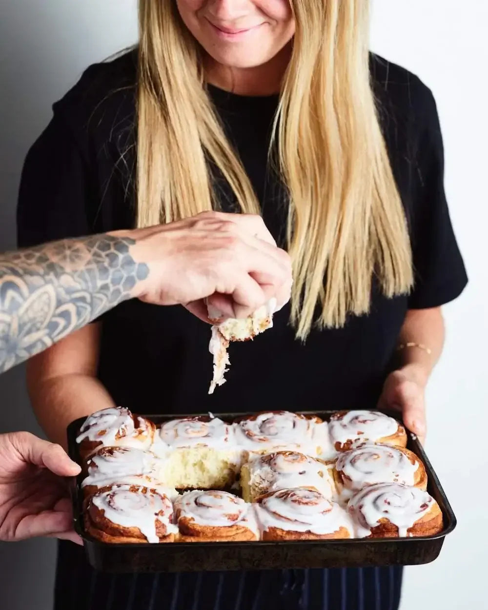 Person holding a tray of iced cinnamon rolls while another hand pulls one apart.