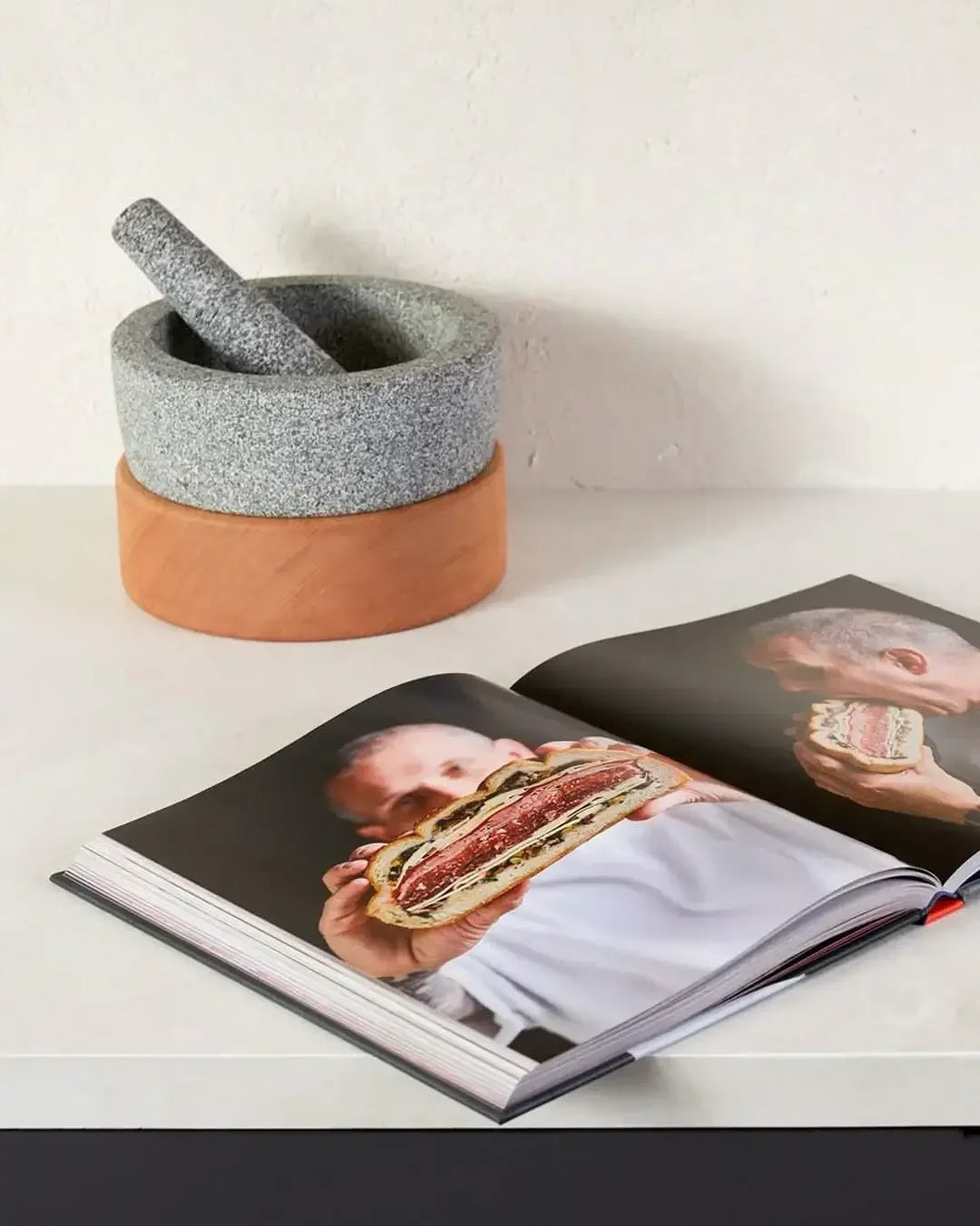 Mortar and pestle beside open cookbook with chef holding gourmet sandwich