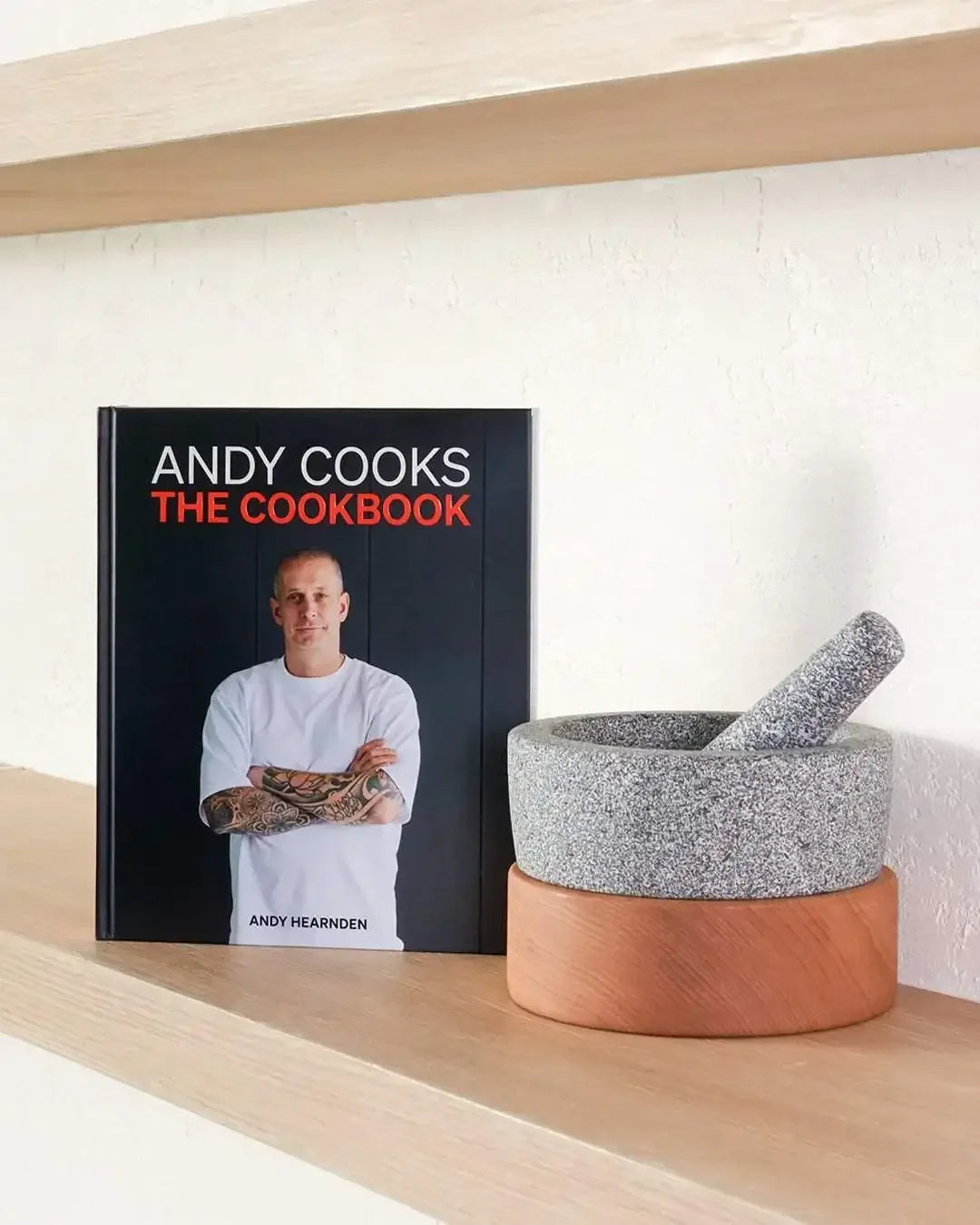 Andy Cooks cookbook standing next to a stone mortar and pestle on a wooden shelf