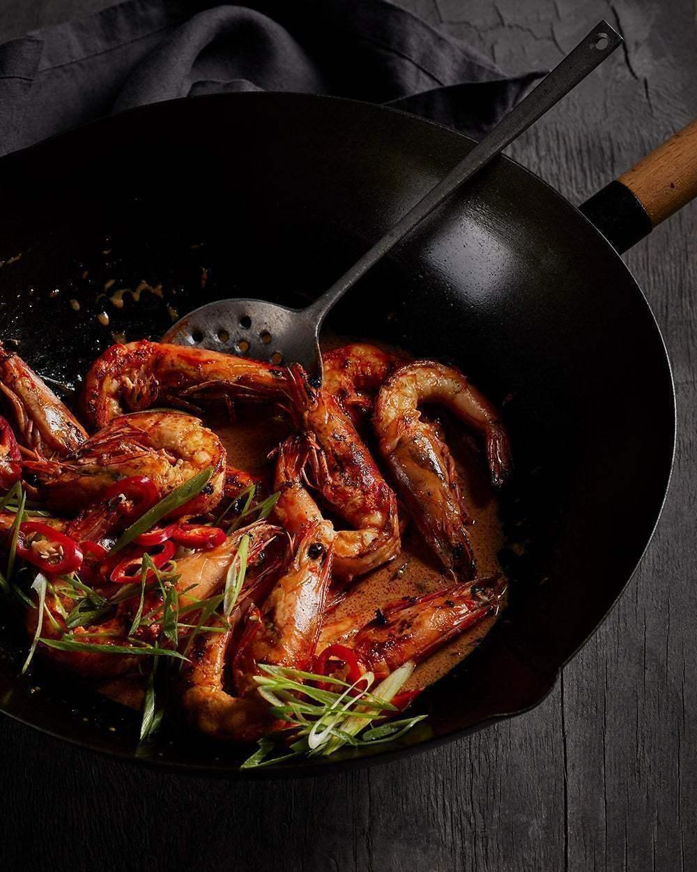 Wok with cooked prawns, sliced red chili, and green onions on a dark wood surface