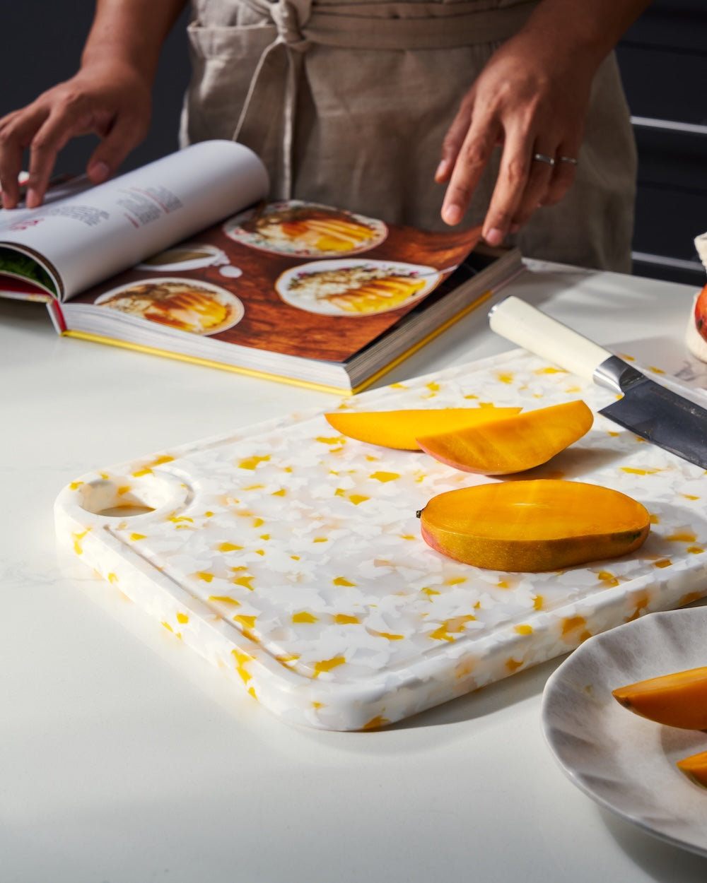 Recycled terrazzo chopping board with sliced mango, cookbook, and kitchen knife on white countertop.