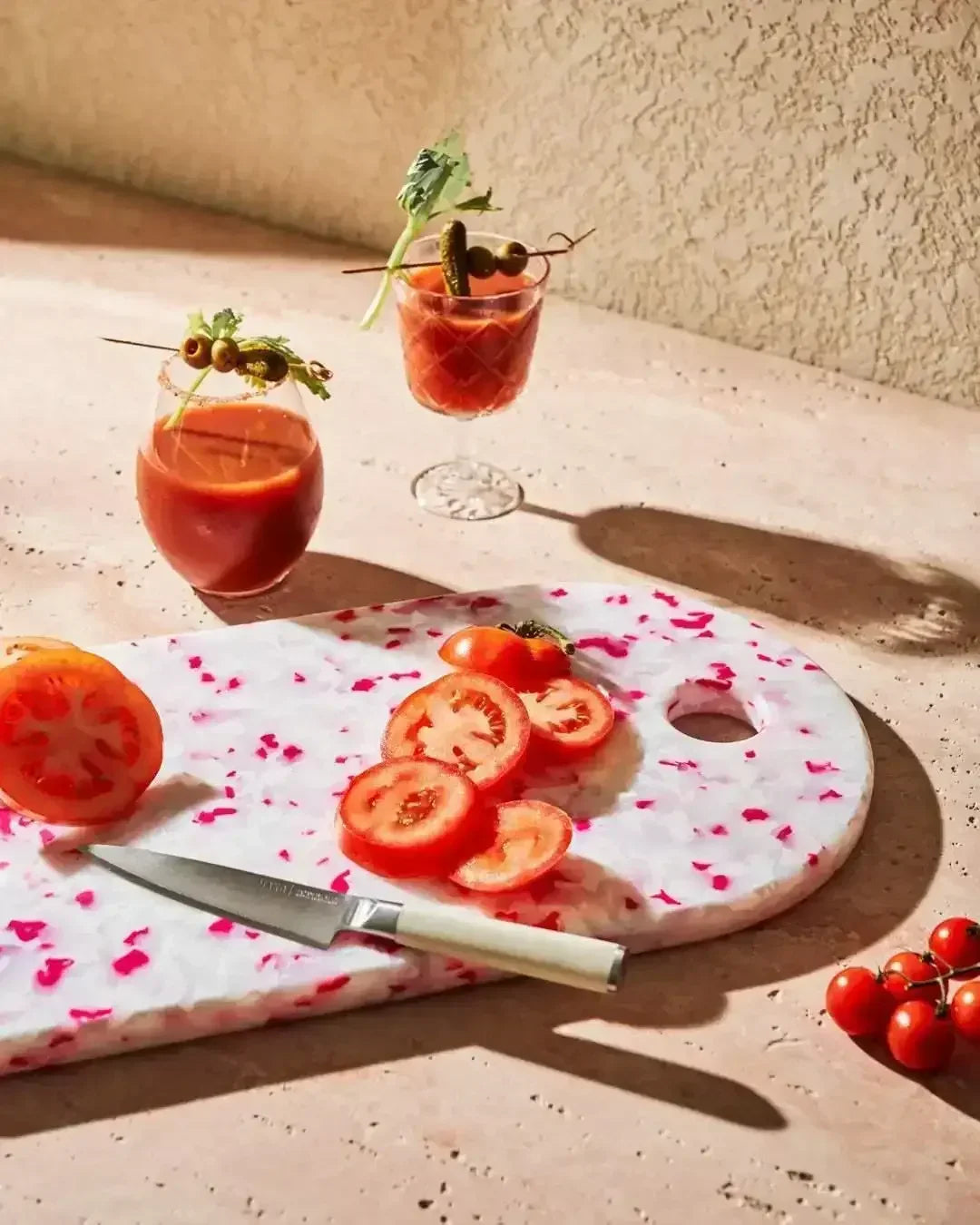 Pink speckled recycled chopping board with sliced tomatoes, kitchen knife, and cocktails nearby.