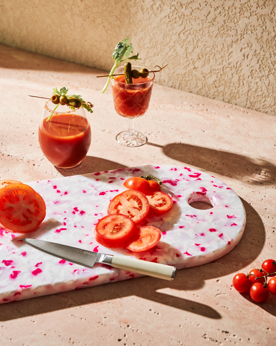 Pink speckled recycled chopping board with sliced tomatoes, kitchen knife, and cocktails nearby.