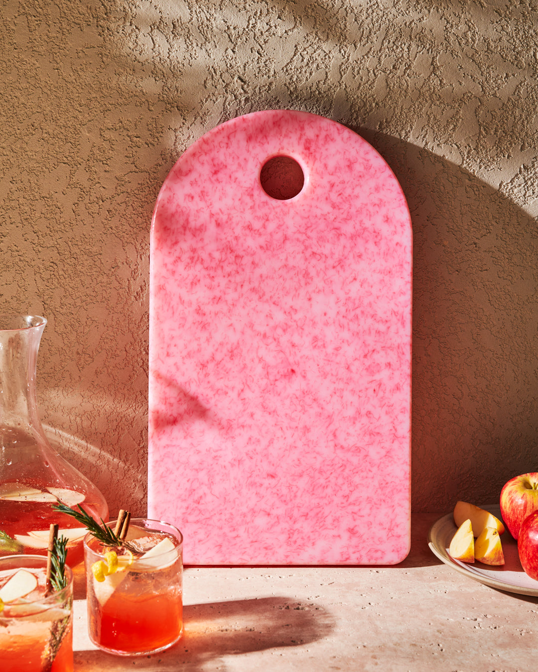 Pink recycled chopping board with marbled pattern, arched top, surrounded by apples and drinks