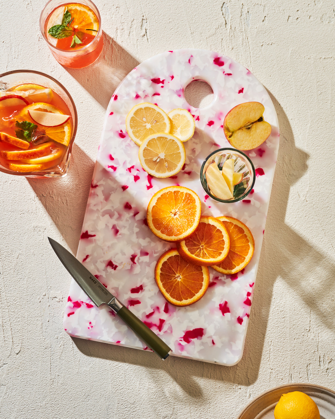 Colorful chopping board with sliced oranges, lemons, apple, knife and glass of fruit on table