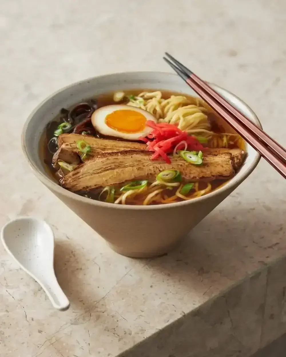 Ceramic ramen bowl with noodles, egg, pork slices, chopsticks, and white spoon on stone surface