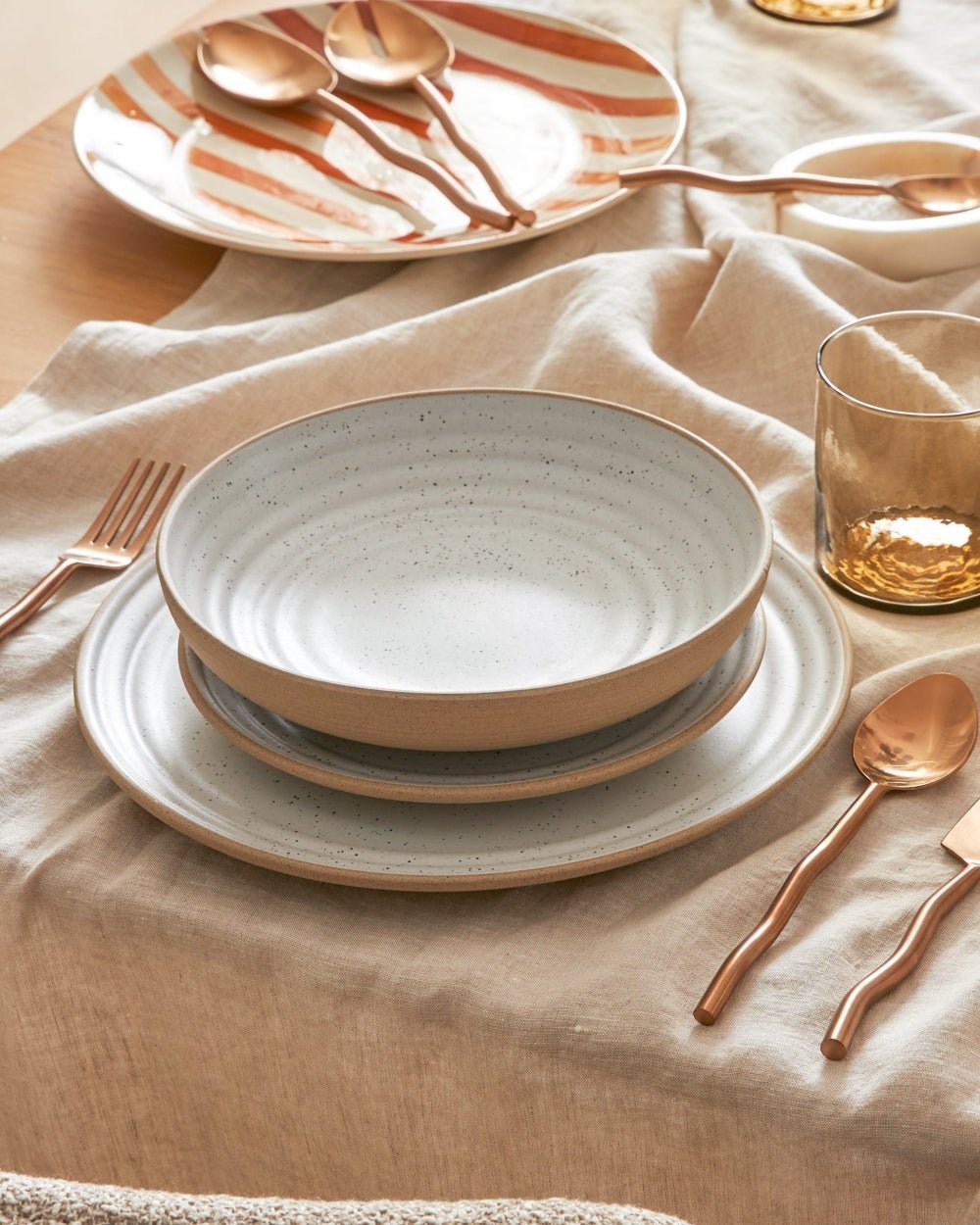 Neutral stoneware dinner plate, bowl, and salad plate set on beige tablecloth with gold cutlery.
