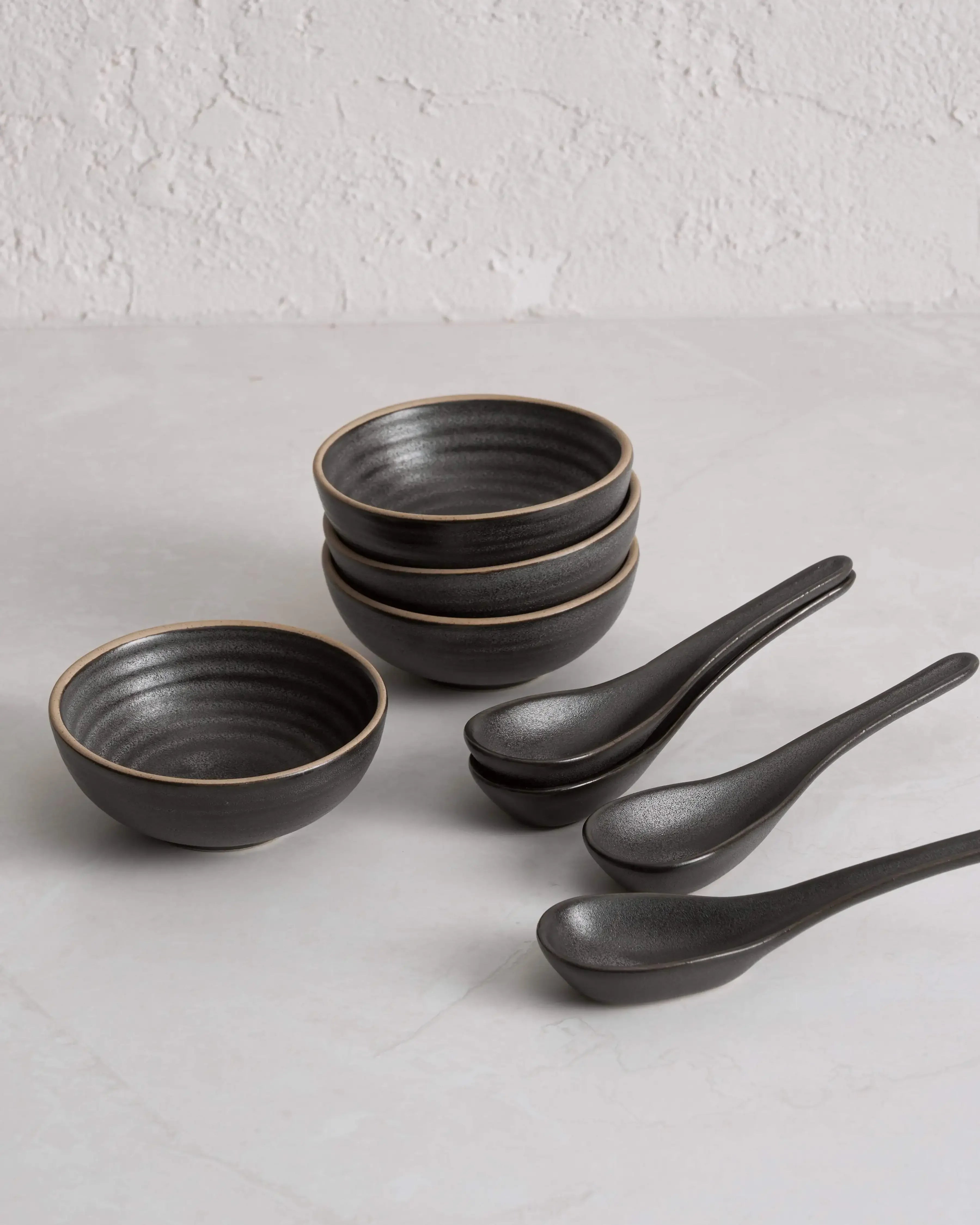 Charcoal stoneware bowls and ceramic soup spoons set on light textured surface