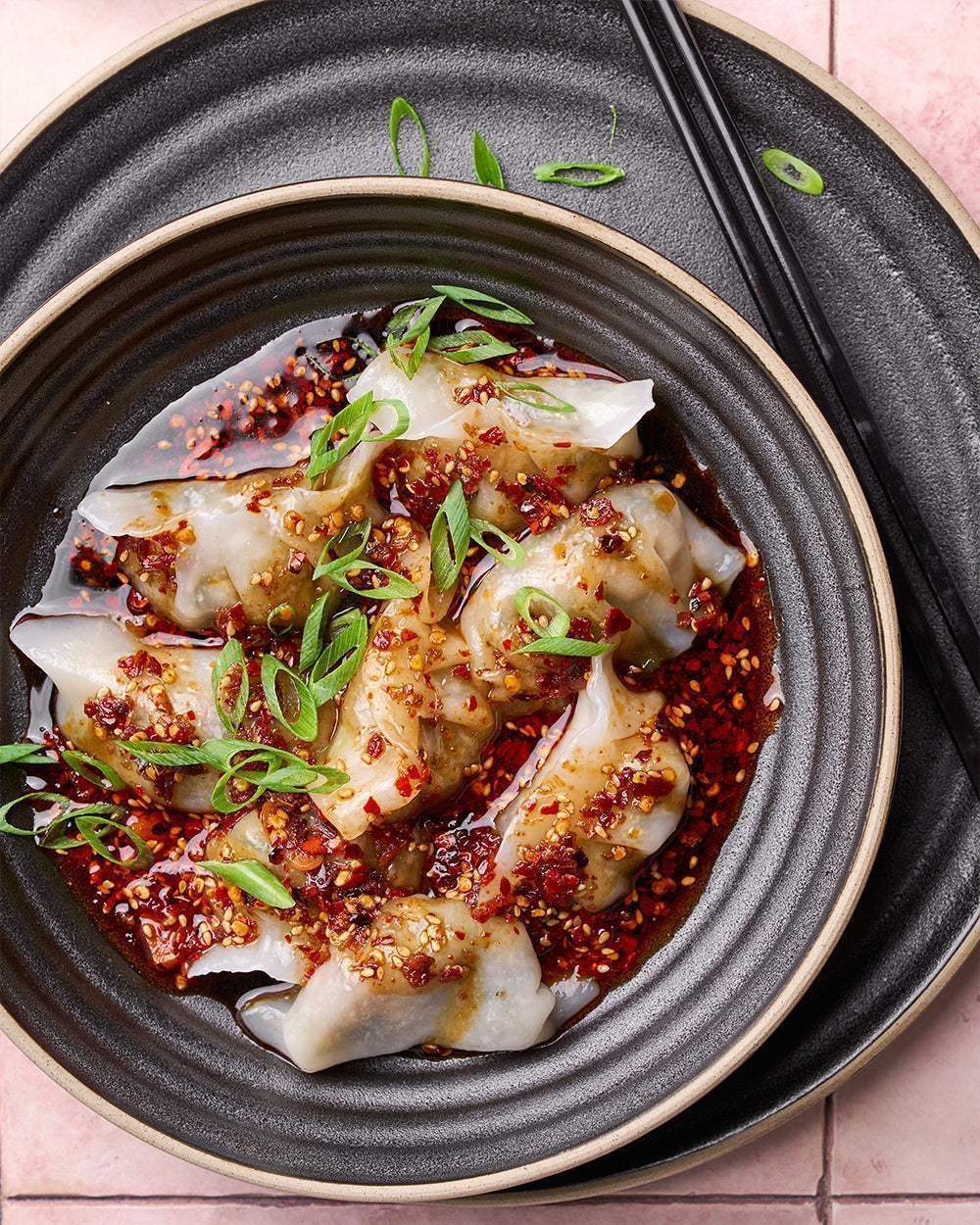 Charcoal Asoke dinnerware plate with dumplings in chili oil garnished with sliced spring onions and black chopsticks