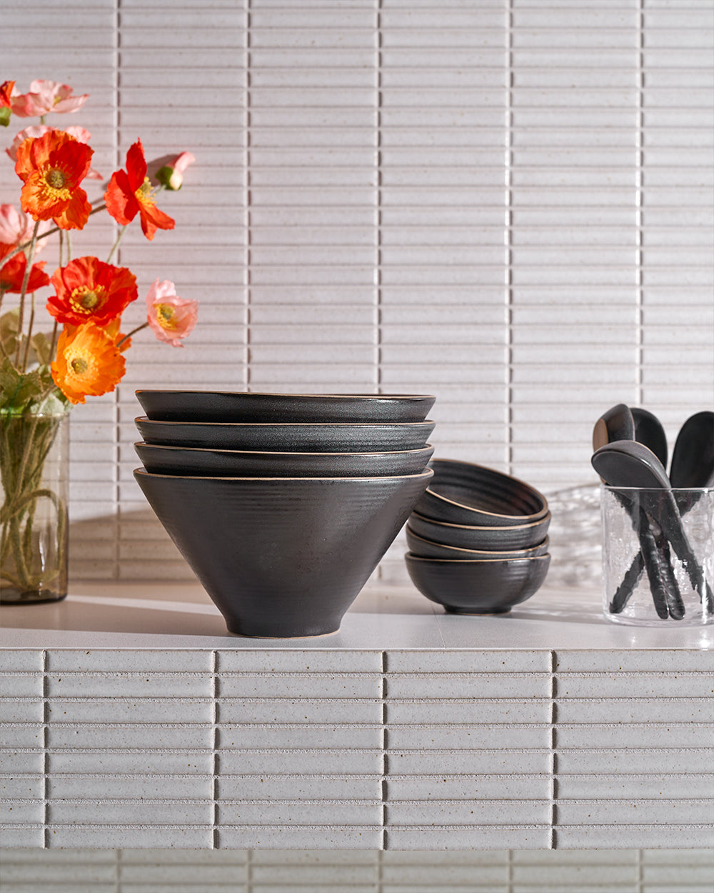 Charcoal ceramic dinnerware set with bowls and plates on a white tiled shelf, flowers in vase