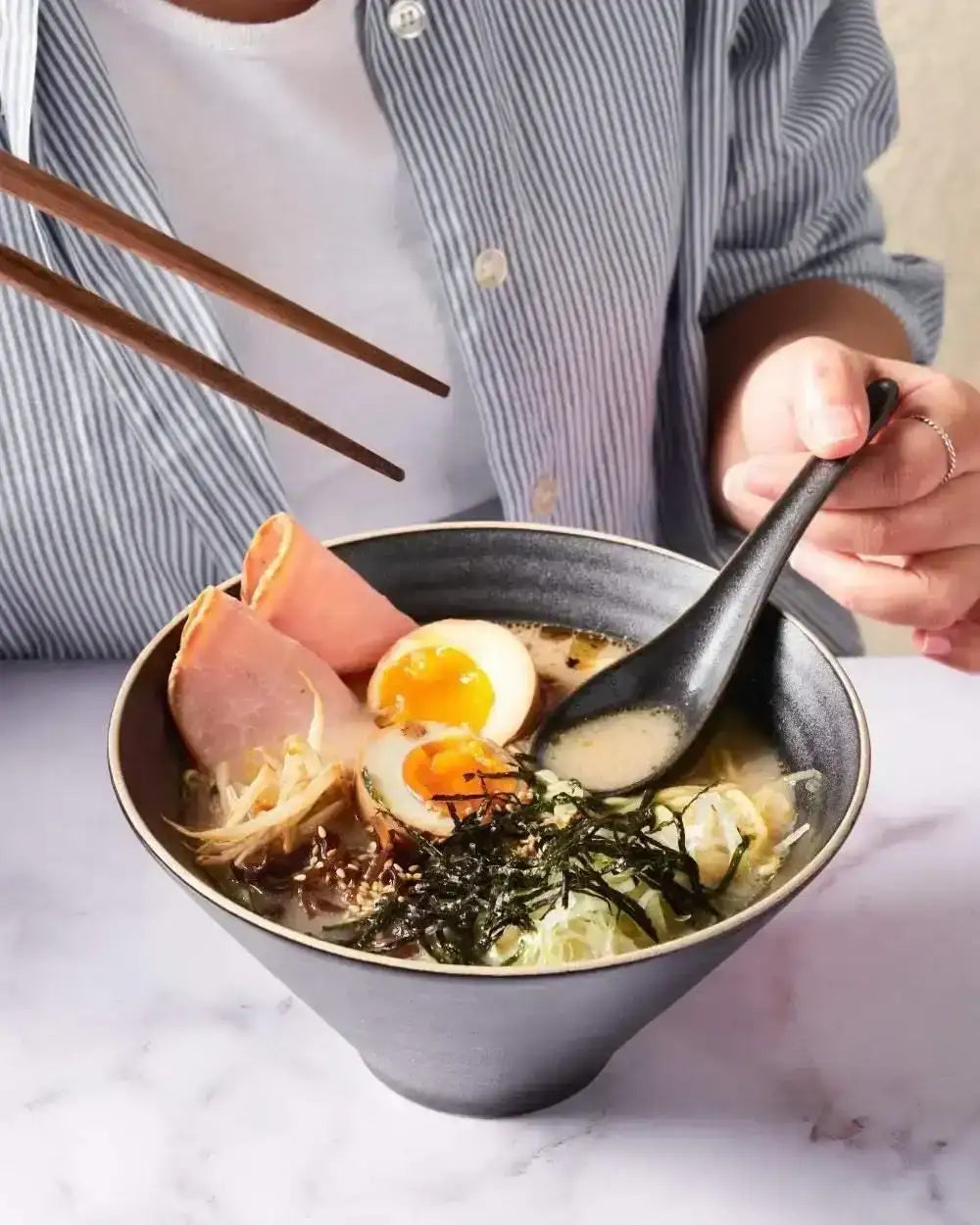 Person eating ramen with chopsticks and black spoon in charcoal bowl by Marion Grasby