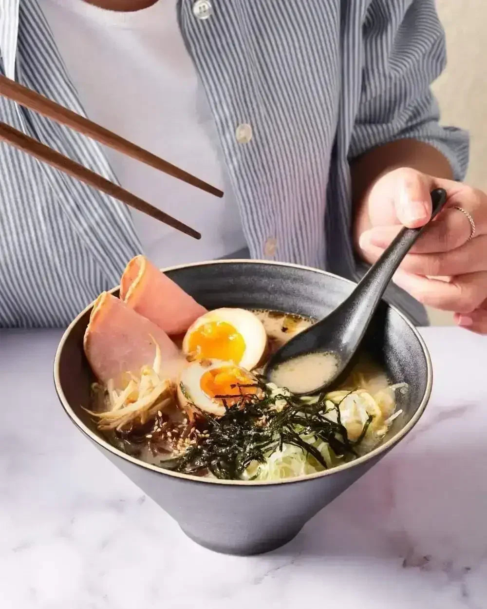 Charcoal ceramic noodle bowl holding ramen with egg, ham, seaweed, and vegetables