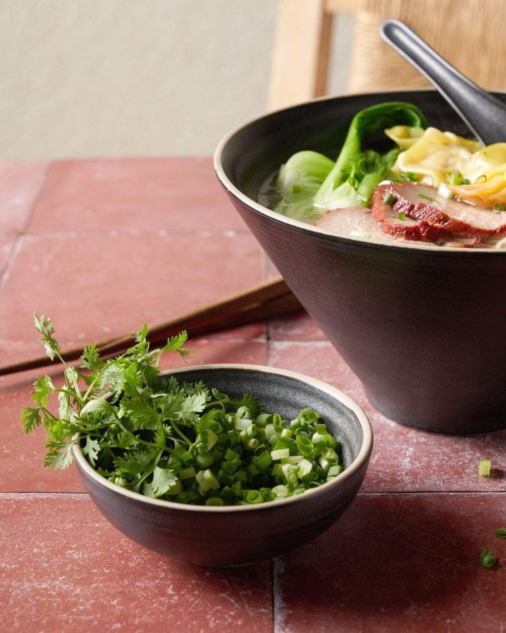 Asoke charcoal ceramic noodle bowl set with soup, vegetables, and herbs on red tiled surface by Marion Grasby