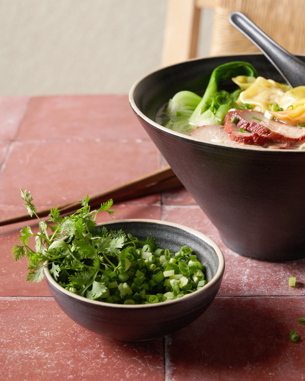 Charcoal ceramic noodle bowl and side dish bowl with fresh herbs and chopped scallions on a red tiled table.