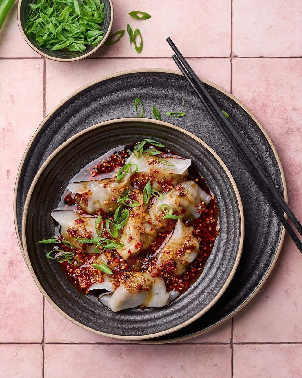 Asoke charcoal dinnerware with Asian dumplings in chili sauce, sprinkled with green onions, black chopsticks on pink tiles