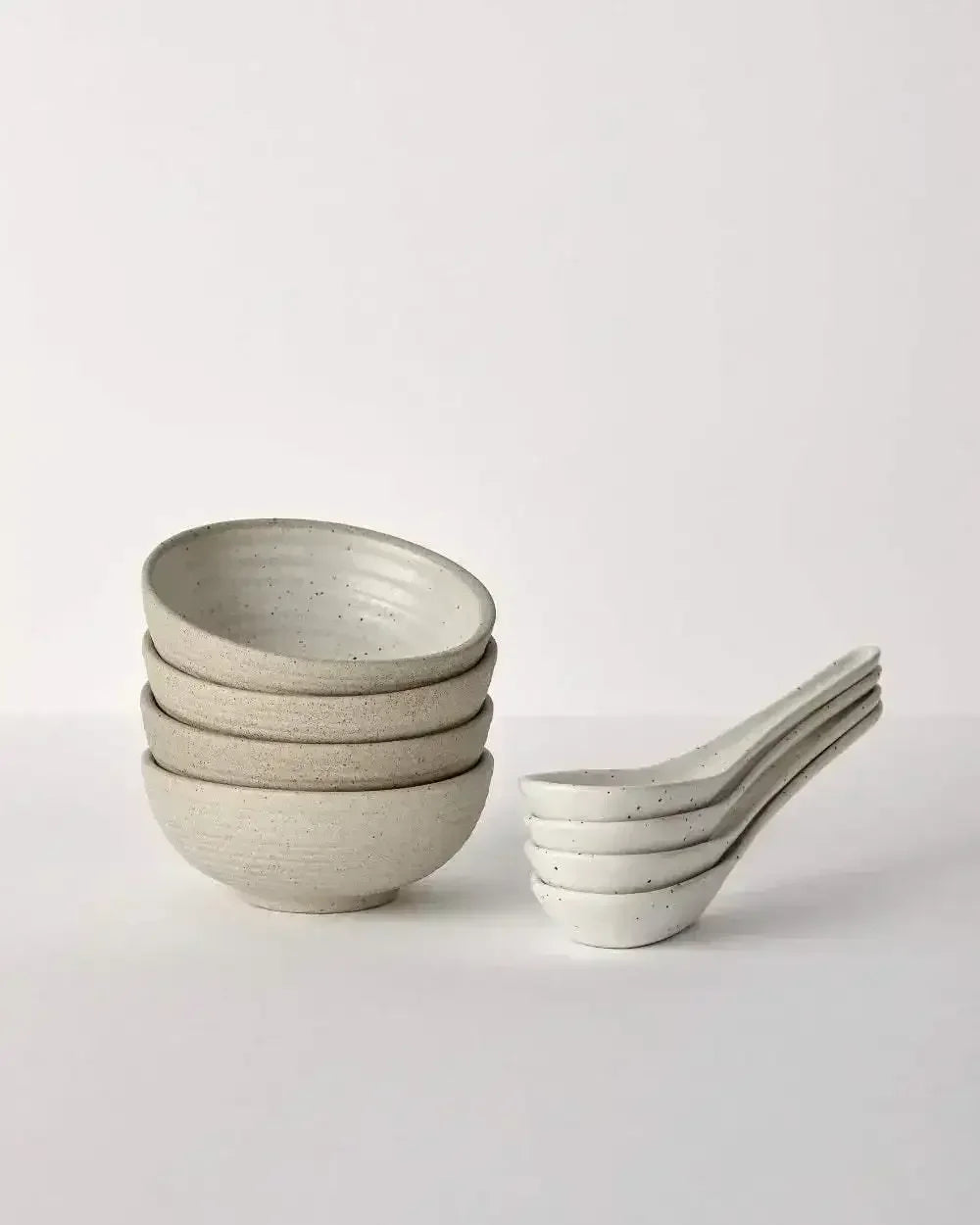 Stacked stoneware bowls and spoons with speckled finish on a white background, dinnerware set