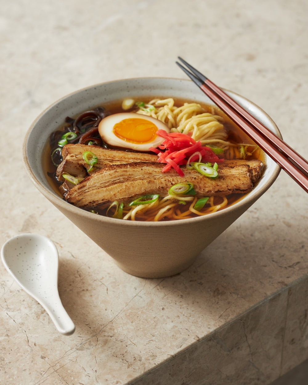 Ceramic noodle bowl filled with ramen, topped with egg, pork, scallions, and chopsticks.