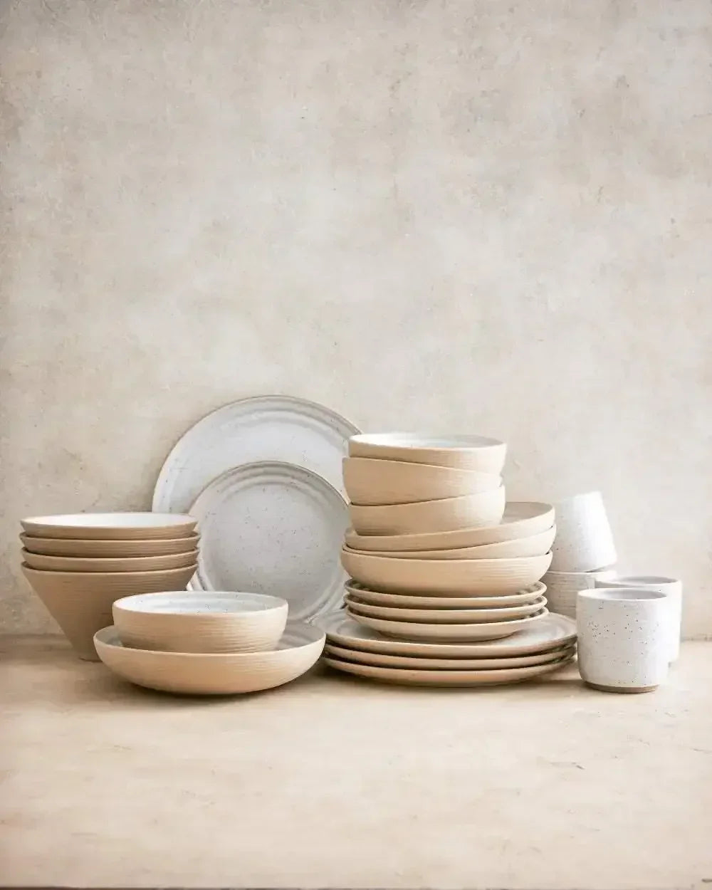 Stacked beige noodle bowls, plates, and speckled ceramic cups on a neutral surface