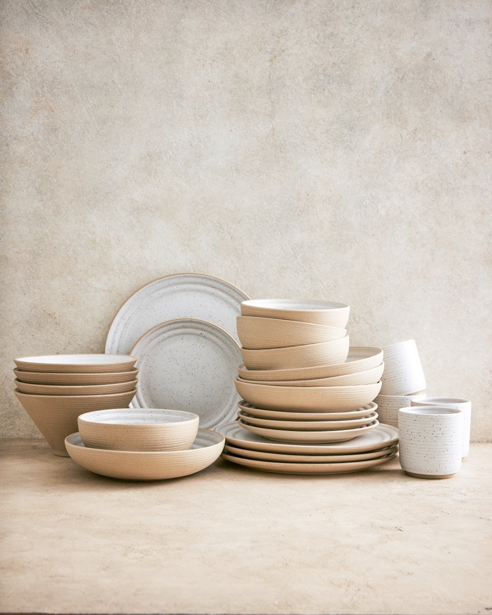 Stacked beige noodle bowls, plates, and speckled ceramic cups on a neutral surface