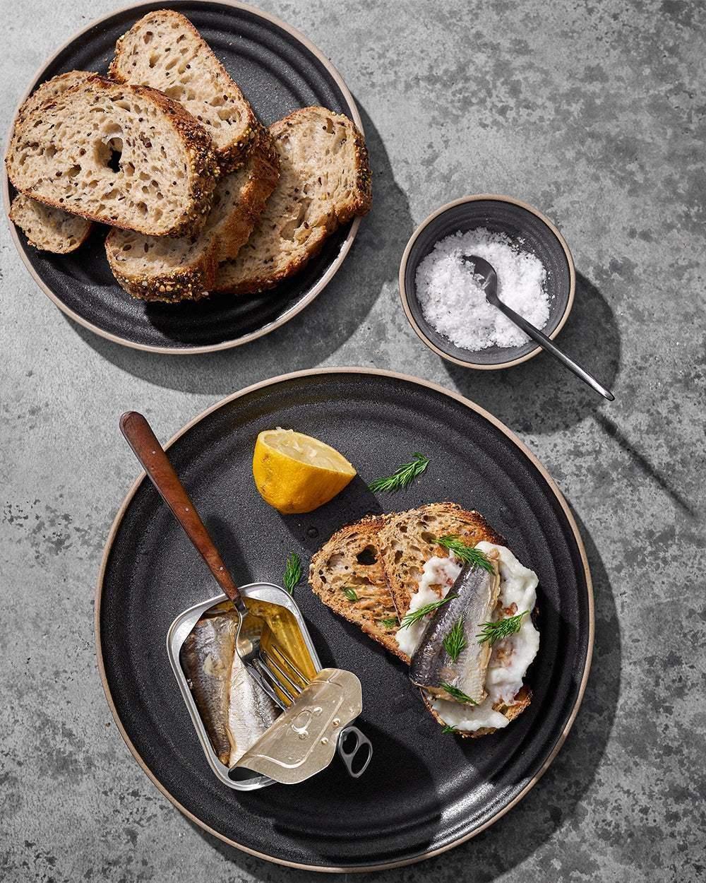 Charcoal ceramic side plates with seeded bread, sardines, lemon wedge, and salt on a stone surface