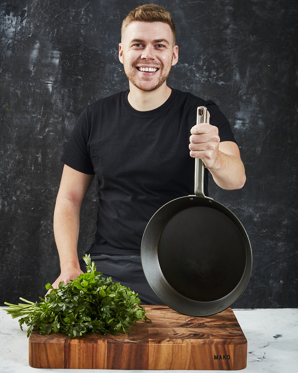 Smiling man holding nonstick frying pan beside fresh herbs on wooden cutting board