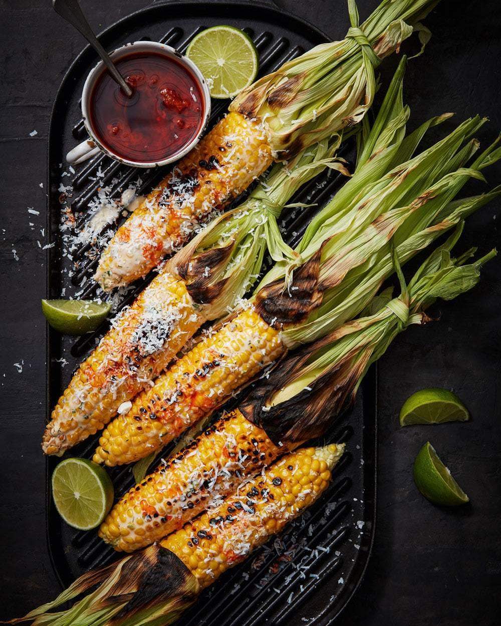 Grilled corn on the cob with charred husks, lime wedges, and a cup of red sauce on a black grill pan