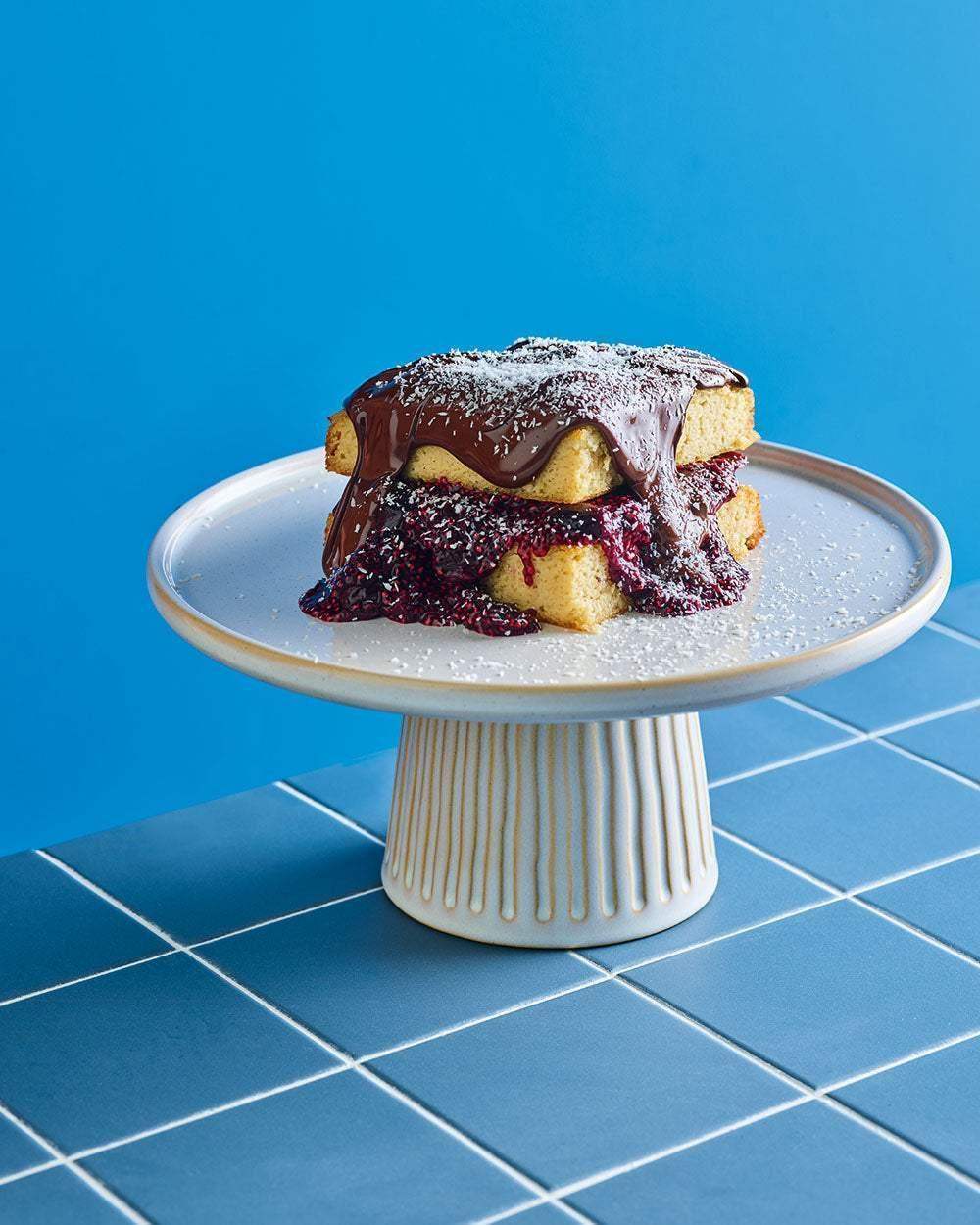 Two-layer sponge cake with berry sauce and chocolate glaze on white cake stand, blue tiled table background
