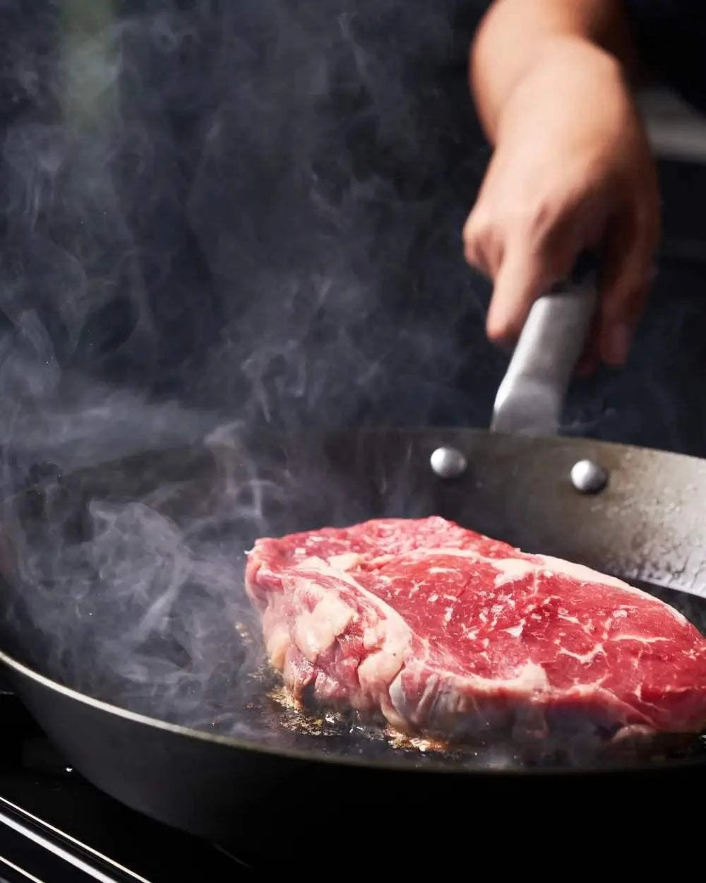 Raw steak searing in a black steel pan with visible smoke, hand holding pan handle