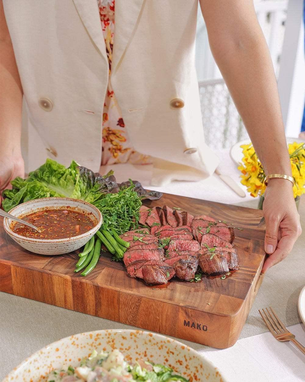 Wooden chopping board with sliced steak, fresh greens, and sauce, held on a dining table
